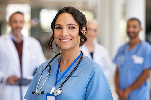 Портрет улыбающейся молодой медсестры в униформе, улыбающейся на фоне команды здравоохранения. Успешная команда врачей и медсестер улыбается. Красивый и довольный медицинский работник стоит в частной клинике и с гордостью смотрит в камеру.
